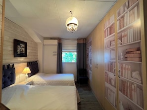 Une chambre d’hôtel avec deux lits, un lustre, une bibliothèque et une fenêtre avec des rideaux.