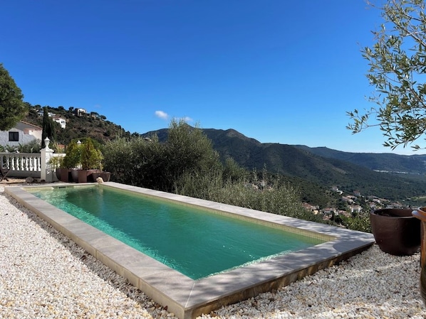 Une piscine rectangulaire bordée de galets, elle-même entourée de petits cailloux blancs, le tout se fondant dans un paysage montagneux aux couleurs vives, sous un ciel bleu dégagé.