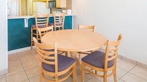 Une table ronde en bois, entourée de quatre chaises, chacune dotée d’un coussin. La pièce est dotée d’un sol carrelé et comprend un coin cuisine avec des placards jaunes et un plan de travail blanc.
