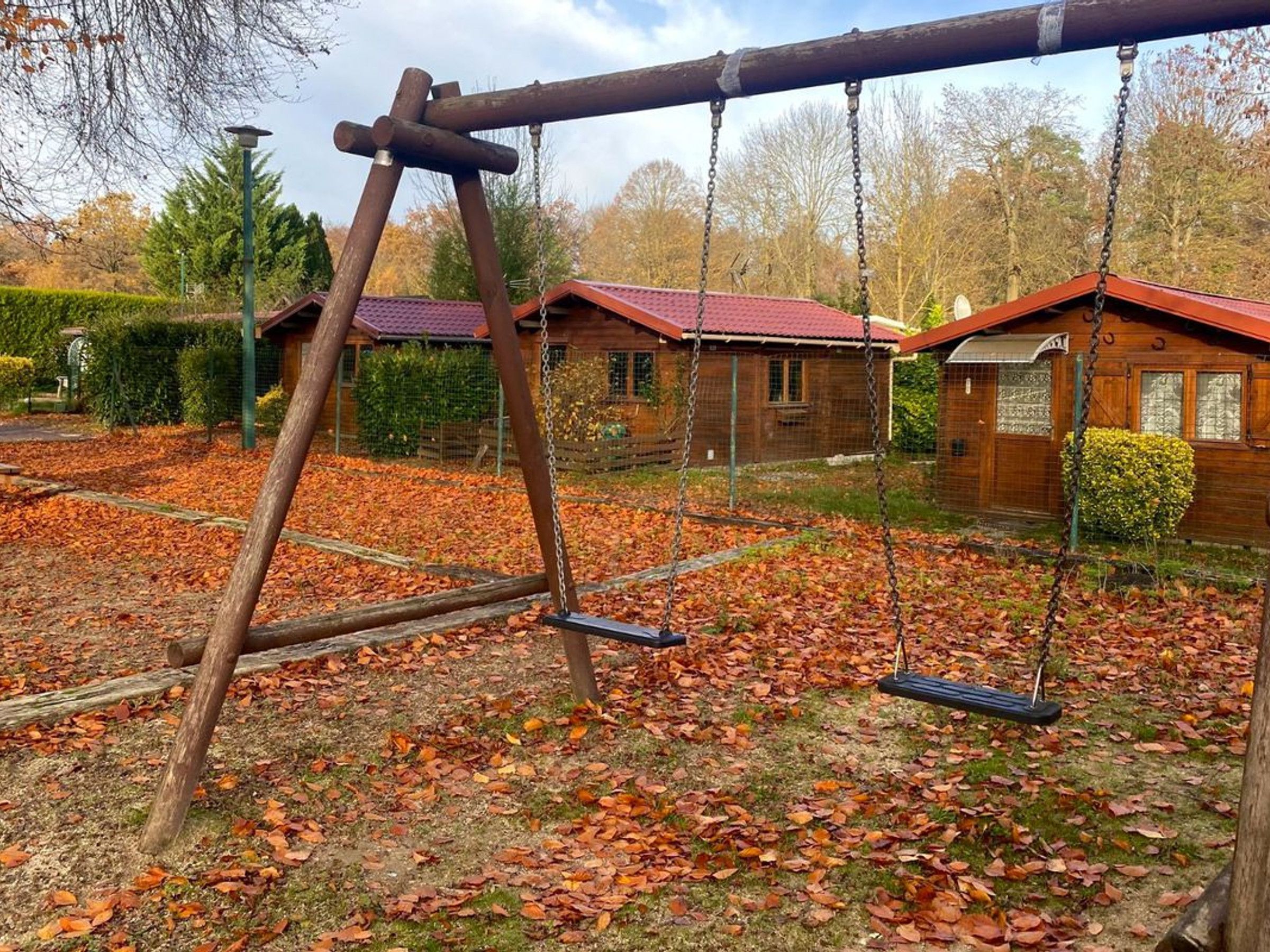 Un ensemble de balançoires en bois dans un parc, avec un sol recouvert de feuilles, entouré de bâtiments aux toits rouges.