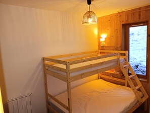 Quarto com beliche, teto de madeira, luminária na parede e janela com vista para uma paisagem nevada.
