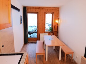 Interior em madeira com uma mesa de jantar e bancos, um radiador, uma janela com vista para rochas cobertas de neve e uma porta.