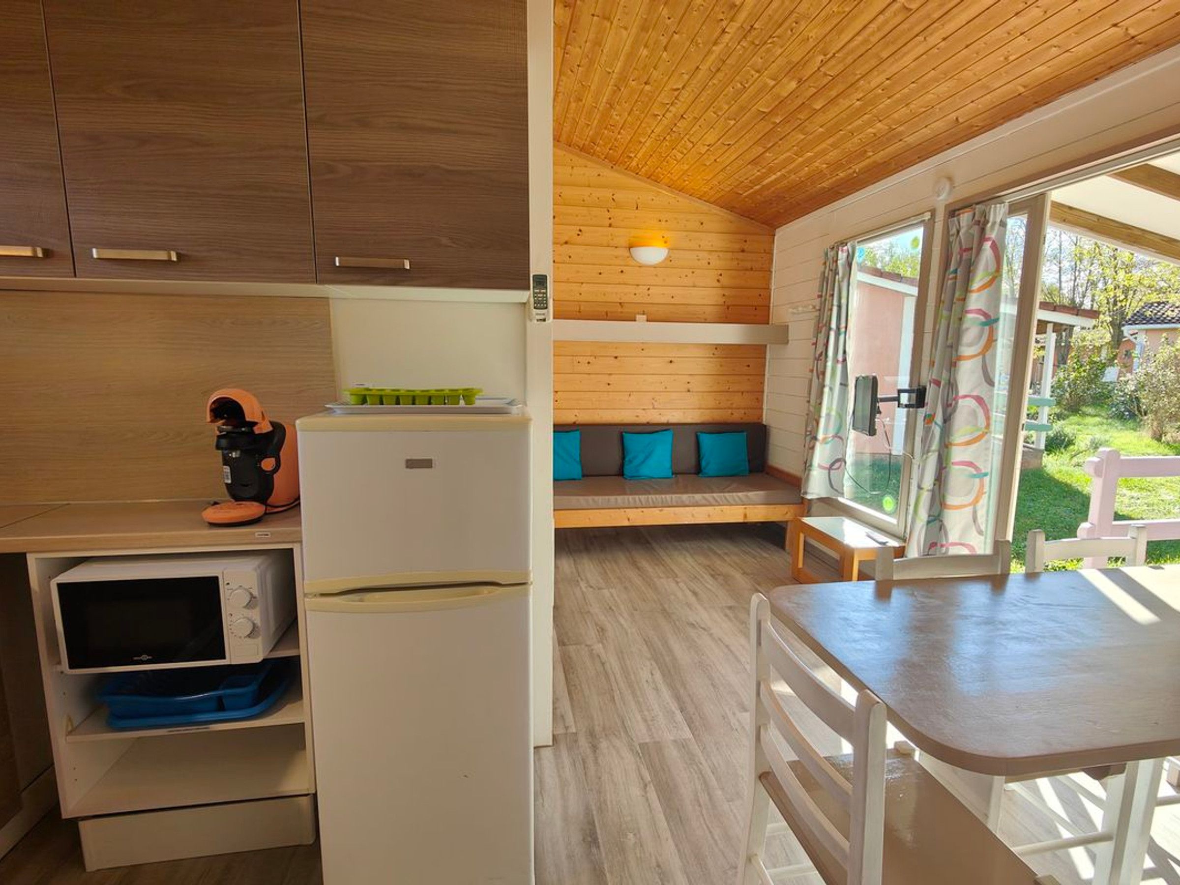 Cocina compacta con microondas, refrigerador y un lavamanos pequeño. En la zona de estar contigua, hay un banco de madera con cojines.