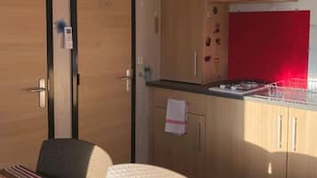 Une petite pièce comprenant un lit, une petite table, un coin cuisine avec un plan de travail rouge, et une porte sur laquelle est accrochée une serviette.