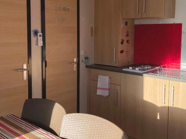A compact room with a bed, a small table, a kitchenette with a red backsplash, and a door with a towel hanging on it. - Mobile home, AC, pets welcome - 2 guests (Angicourt)