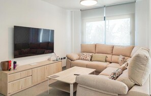 A modern living room with a flat-screen TV, a beige sofa, a wooden coffee table, and a window with blinds.
