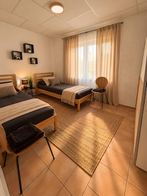 Een slaapkamer met twee bedden, een stoel, een nachtkastje met een lamp en een raam met gordijnen.