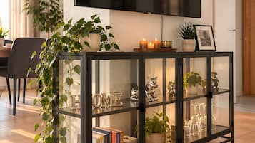 A modern living room with a glass-fronted console table, a flat-screen TV mounted on the wall, and a potted plant on the table.