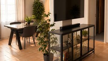 A modern living room with a flat-screen TV on a black console, a dining table with chairs, and a potted plant.