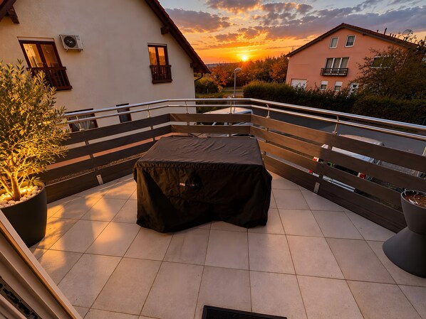 Een terras buiten met een overdekte tafel en stoelen, een bloempot en een hekwerk met uitzicht op een woonwijk tijdens de zonsondergang.