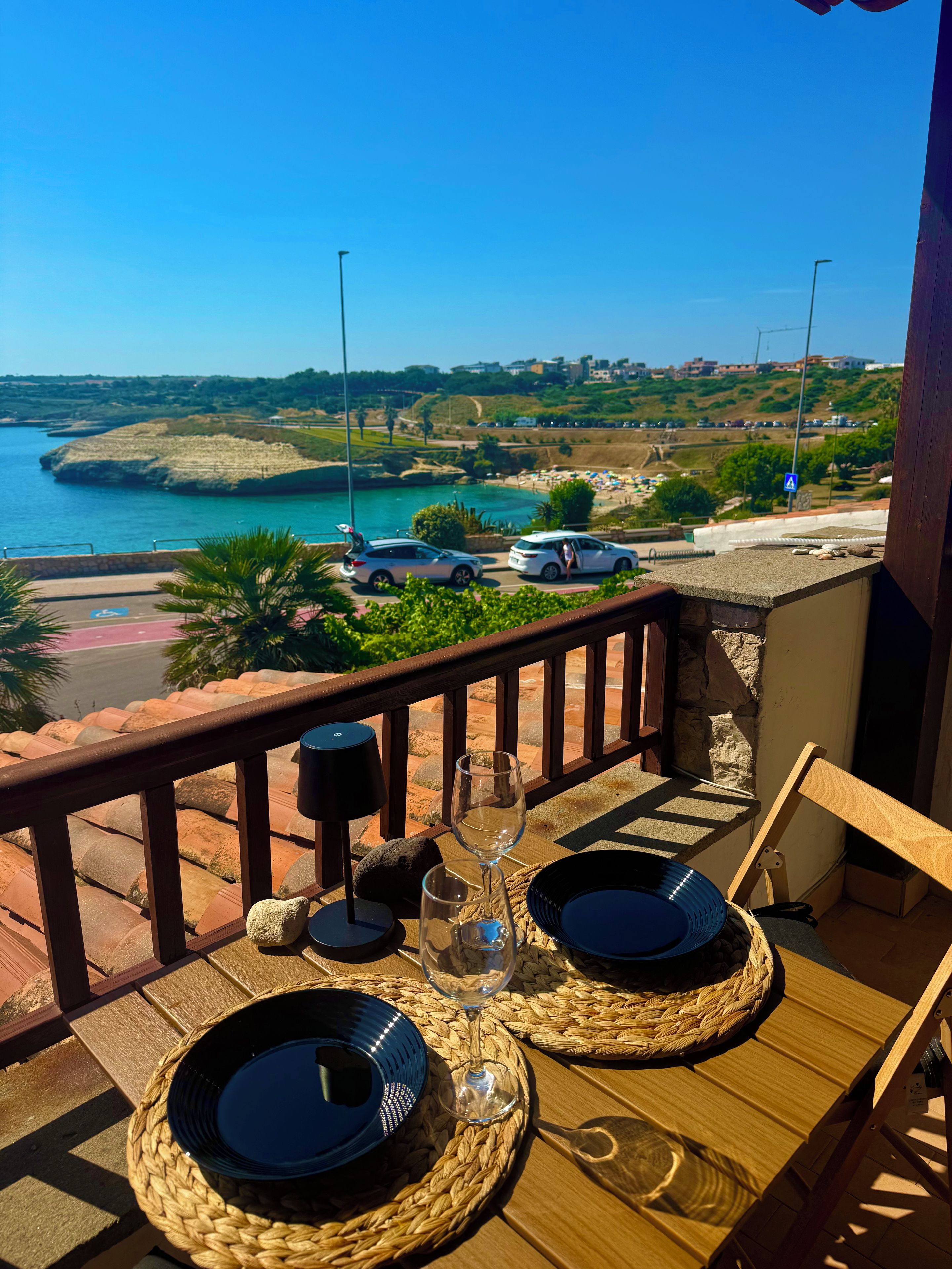 Un balcone con un tavolo apparecchiato per due, con vista su una zona costiera, auto parcheggiate e il mare.