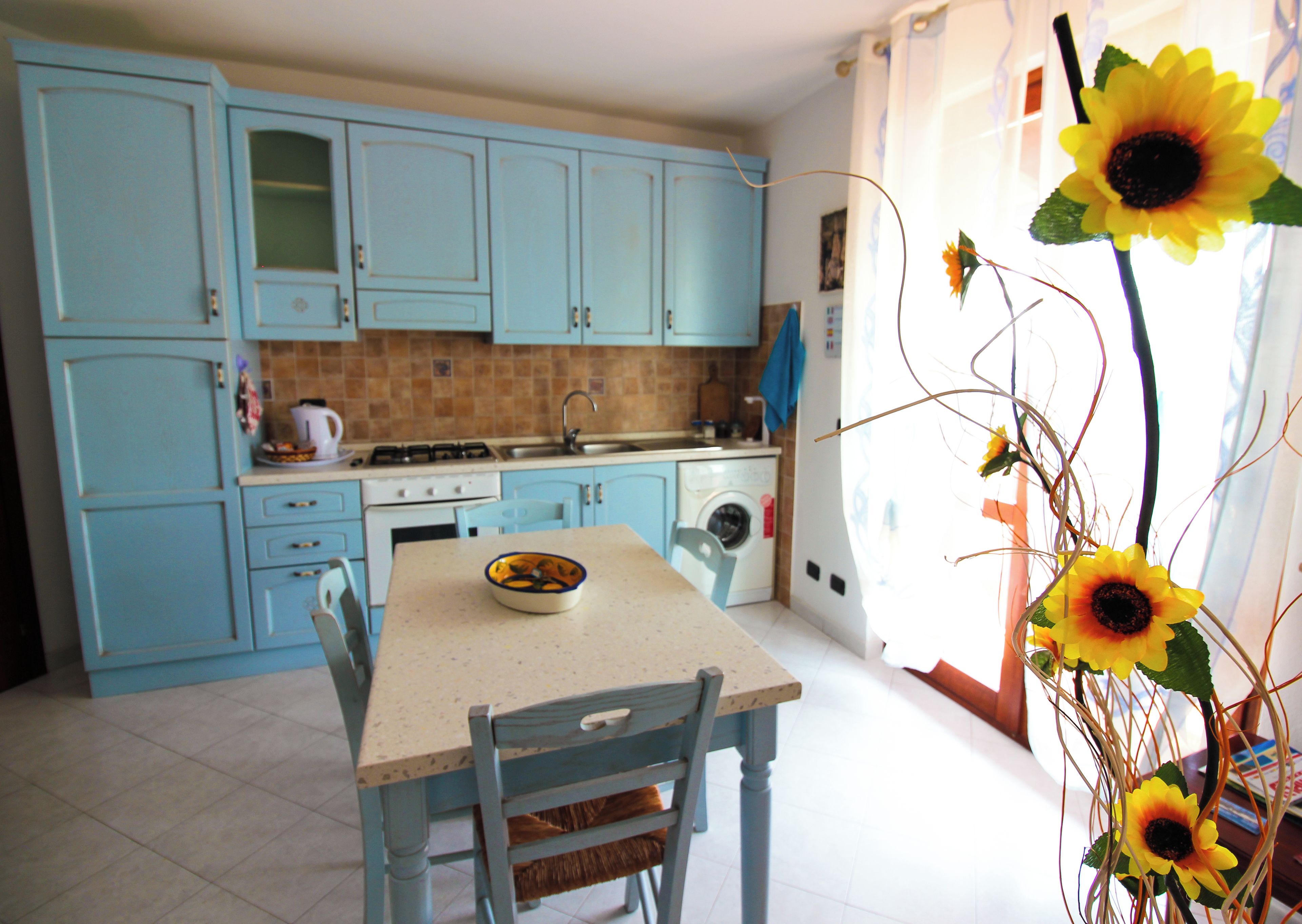 Una cucina con mobili azzurri, un tavolo da pranzo con sedie e un vaso con girasoli.