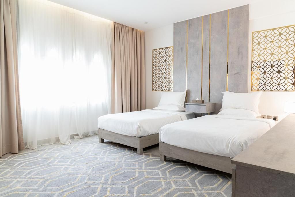 Ett hotellrum med två sängar, ett stort fönster med tunna, genomskinliga gardiner, en väggavskärmare med ett geometriskt mönster och en mönstrad matta.