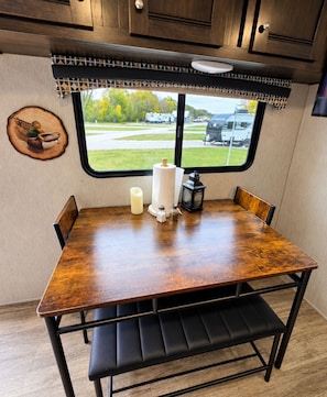 Un comedor con mesa de madera, banca acolchada negra y ventana con vista a un área verde y vehículos.