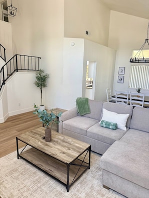 Amplia sala de estar con escalera, un sofá gris, una mesa de centro de madera y una planta en maceta.