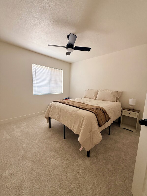 Un dormitorio con una cama, una mesita de noche, una lámpara, un ventilador de techo y una ventana con persianas.