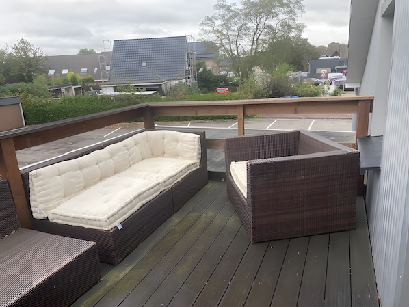A deck with a sofa and two chairs, a parking lot in the background, and a building with a flat roof. - Quiet self-contained apartment with terrace and parking (Heide)