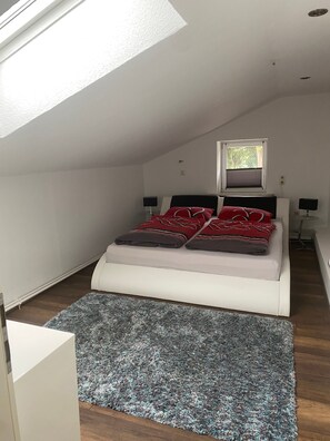 A bedroom with a skylight, a bed with red and grey bedding, a grey rug, and a small window. - Quiet self-contained apartment with terrace and parking (Heide)