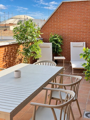 Une terrasse extérieure avec une table et des chaises, entourée de plantes en pot et d’un mur en briques.