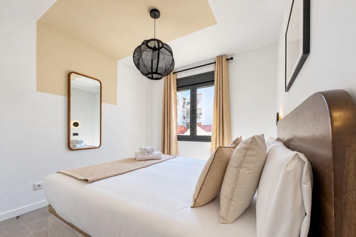 Une chambre à coucher avec un lit, un miroir, un luminaire suspendu et une fenêtre avec des rideaux.