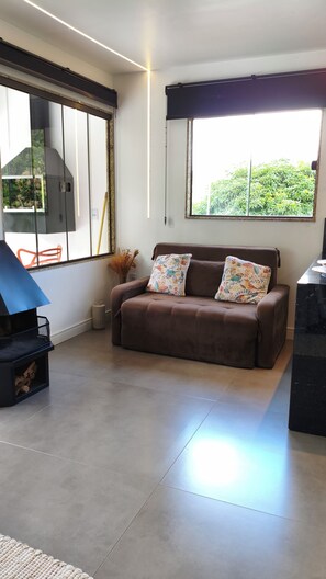 A living room with a brown sofa, two patterned cushions, a large window with a view of greenery, a black grill, and a tiled floor. - cabanas balango (Domingos Martins, Espírito Santo, Brasil)