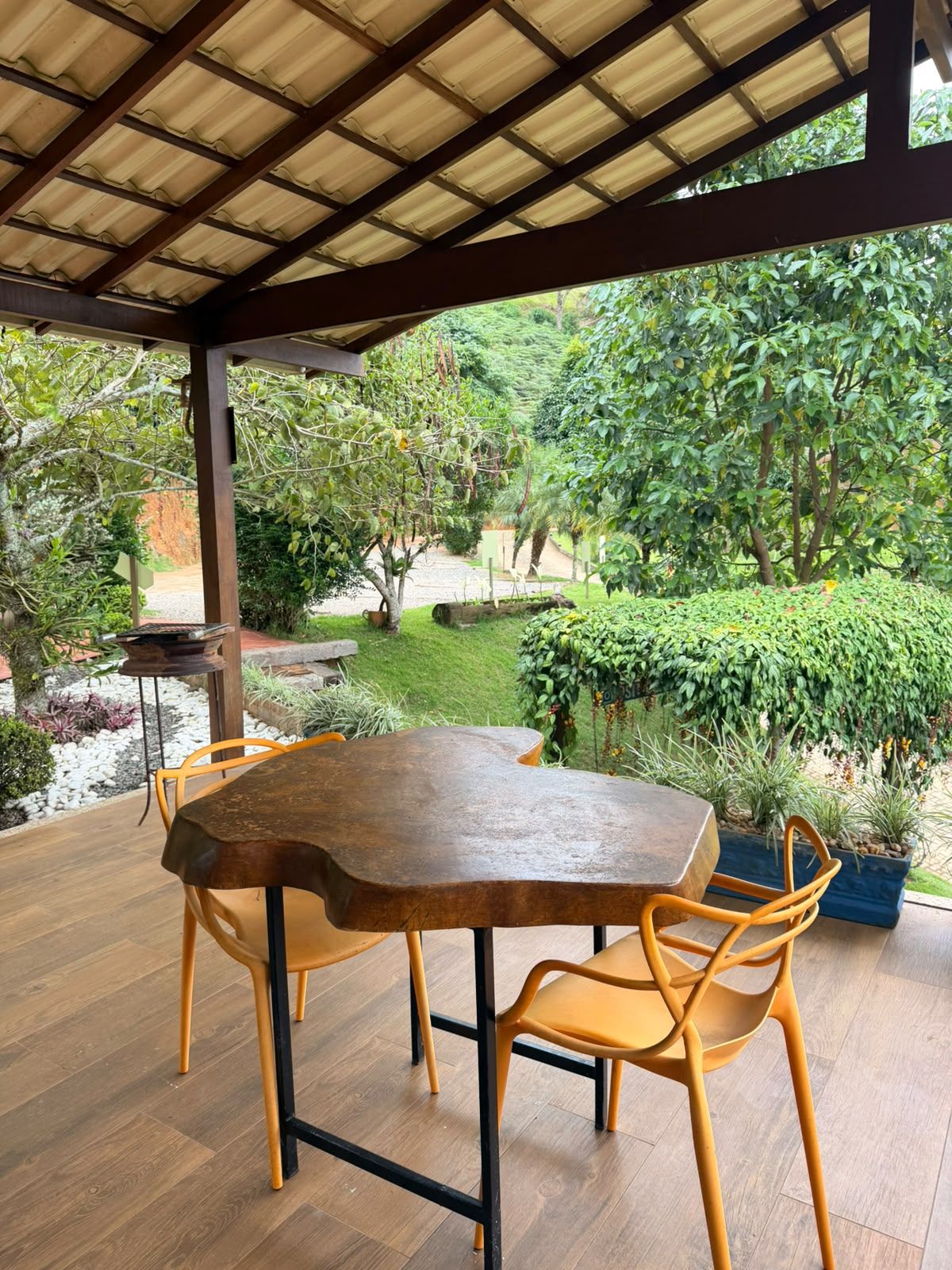 Een terras buiten met een houten tafel en stoelen, omgeven door weelderige begroeiing en uitzicht op een tuin.