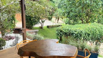 Een terras buiten met een houten tafel en stoelen, omgeven door weelderige begroeiing en uitzicht op een tuin.