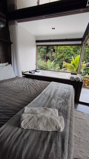 A modern room with a bed, a massage table, and a large window with a view of greenery. - cabanas balango (Domingos Martins, Espírito Santo, Brasil)