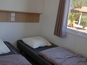 A bedroom with two single beds, a wooden shelf above the beds, and a window with curtains. - Air-conditioned mobile home - 4 guests (Anduze)