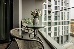 Ein Balkon mit rundem Glastisch, zwei Korbstühlen, einer Blumenvase und Blick auf ein modernes Gebäude.