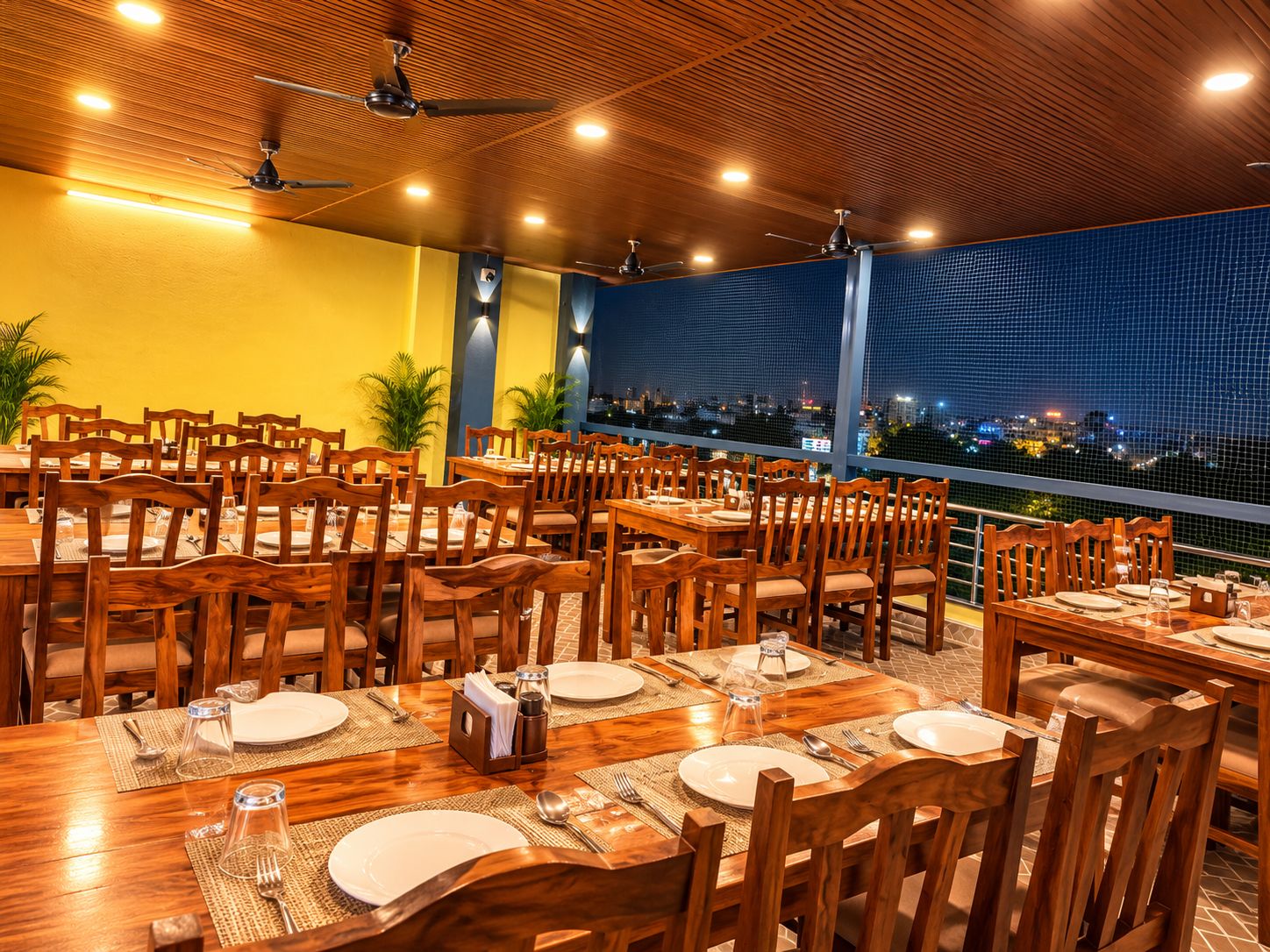 Un restaurante con varias mesas y sillas, ventiladores de techo y vista a la ciudad de noche.