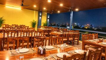 Un restaurante con varias mesas y sillas, ventiladores de techo y vista a la ciudad de noche.