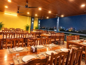 Un restaurante con varias mesas y sillas, ventiladores de techo y vista a la ciudad de noche.