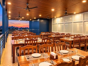 Un restaurante con varias mesas y sillas, ventiladores de techo y amplios ventanales.