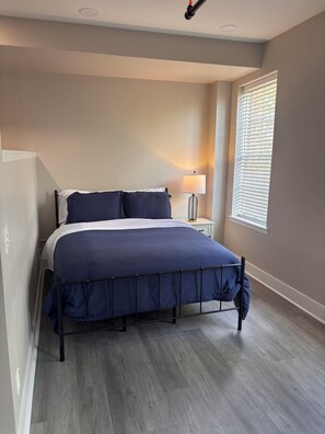 Un dormitorio con cama, lámpara de noche, ventana con persianas y suelo de madera.