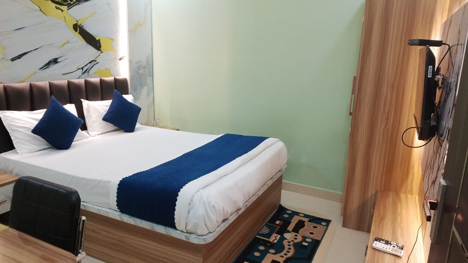A hotel room with a bed, a desk, a chair, a TV, and a wall with a geometric pattern.