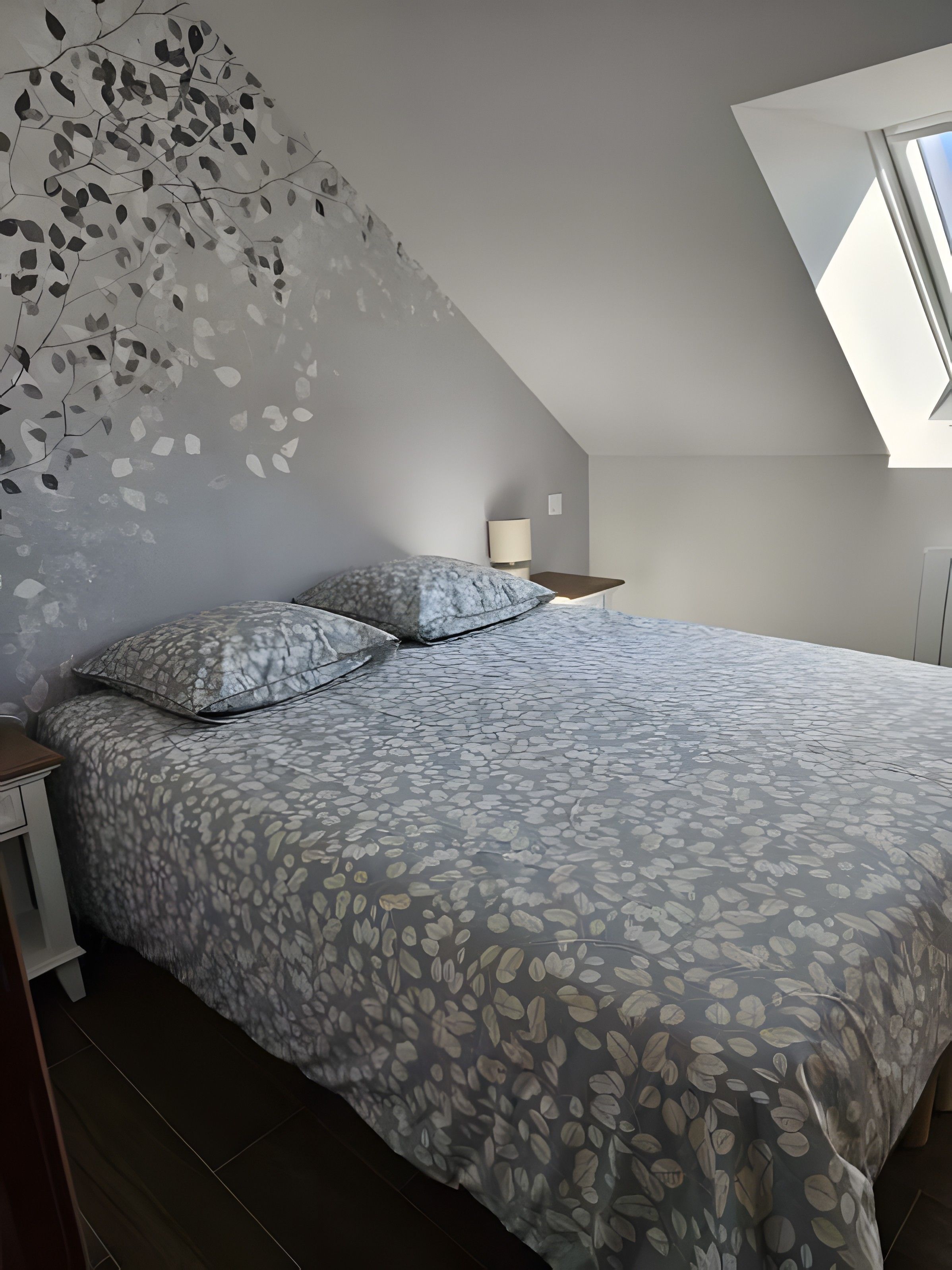 Une chambre à coucher comprenant un lit, une table de chevet, une lampe et un plafond en pente avec un puits de lumière.