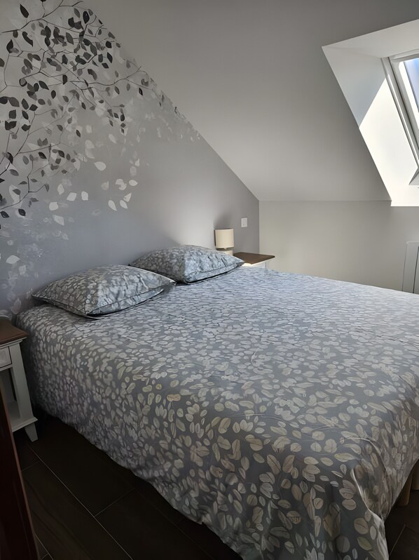 Un dormitorio con cama, mesita de noche, lámpara y techo inclinado con tragaluz.