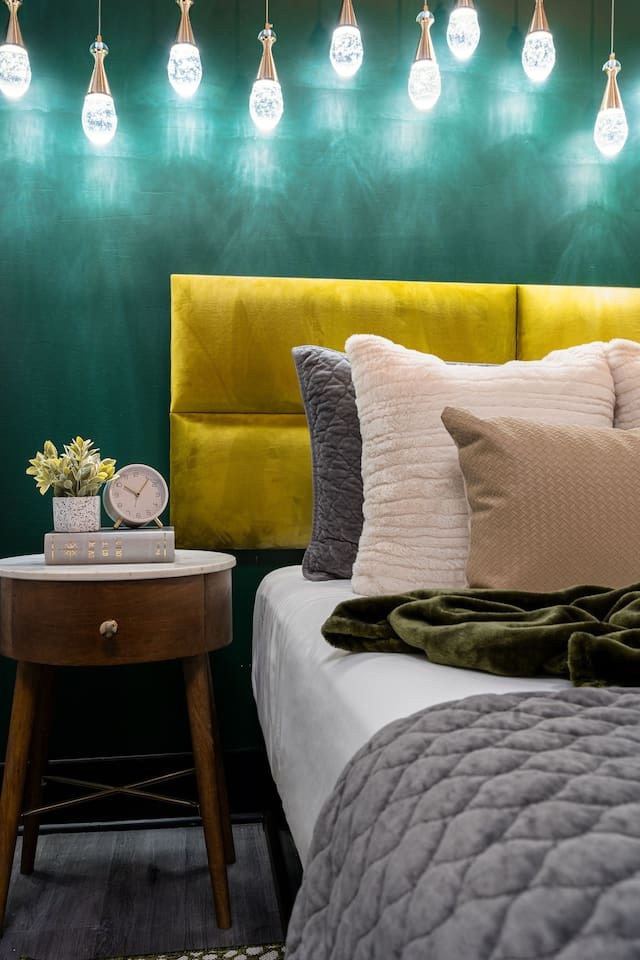 A bedroom with a yellow headboard, a bedside table with a clock and books, and a green blanket on the bed.