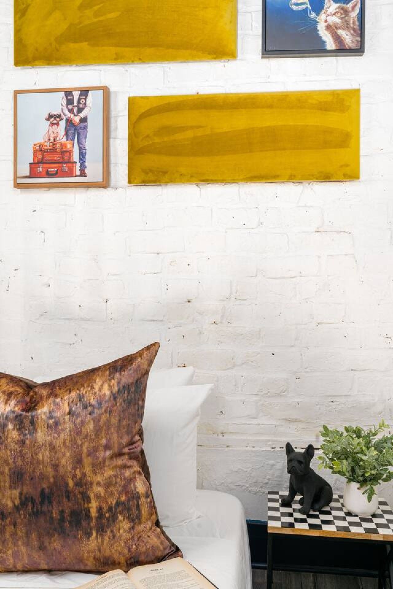 A white brick wall with a large yellow abstract painting, a framed picture of a person with a dog, and a black French Bulldog on a checkered table.