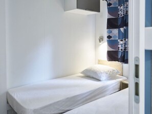 A small, single-bed room with a white bedspread, a white pillow, and a nightstand with a lamp. - Mobile home with terrace - 6 guests (Séez)