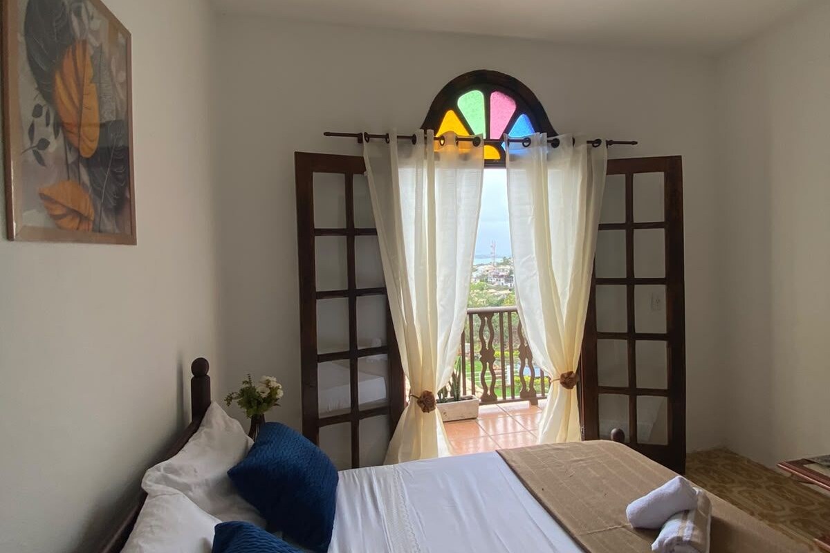 Ein Hotelzimmer mit Bett, Deckenventilator, Fenster mit Vorhängen und einem Bild an der Wand.