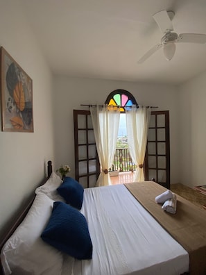 Ein Hotelzimmer mit Bett, Deckenventilator, Fenster mit Vorhängen und einem Bild an der Wand.
