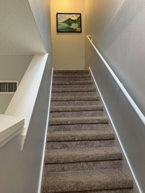 Un escalier recouvert de moquette, avec une main courante blanche, une grille d’aération sur le côté gauche et un tableau encadré au mur.