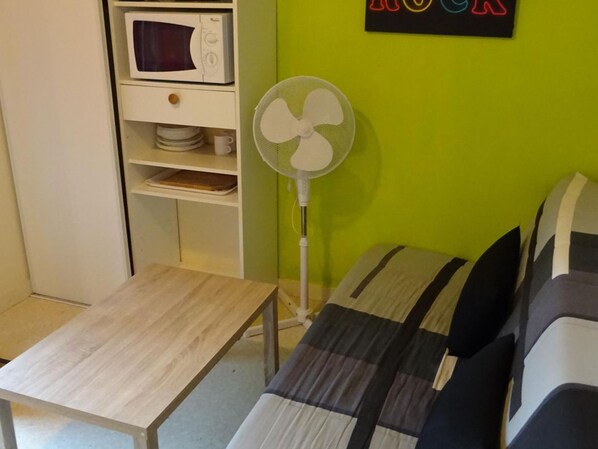 A room with a microwave, a white fan, a small table, and a bed with striped bedding.
