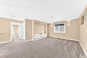 Une pièce spacieuse aux murs de couleur beige, dotée d’un plancher recouvert de moquette, d’une banquette intégrée agrémentée d’objets décoratifs, d’une fenêtre avec des stores et d’un éclairage encastré au plafond.