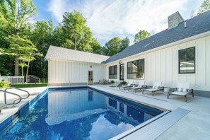 Una piscina con zona de solárium, rodeada por un edificio blanco con ventanas grandes y un porche cubierto.