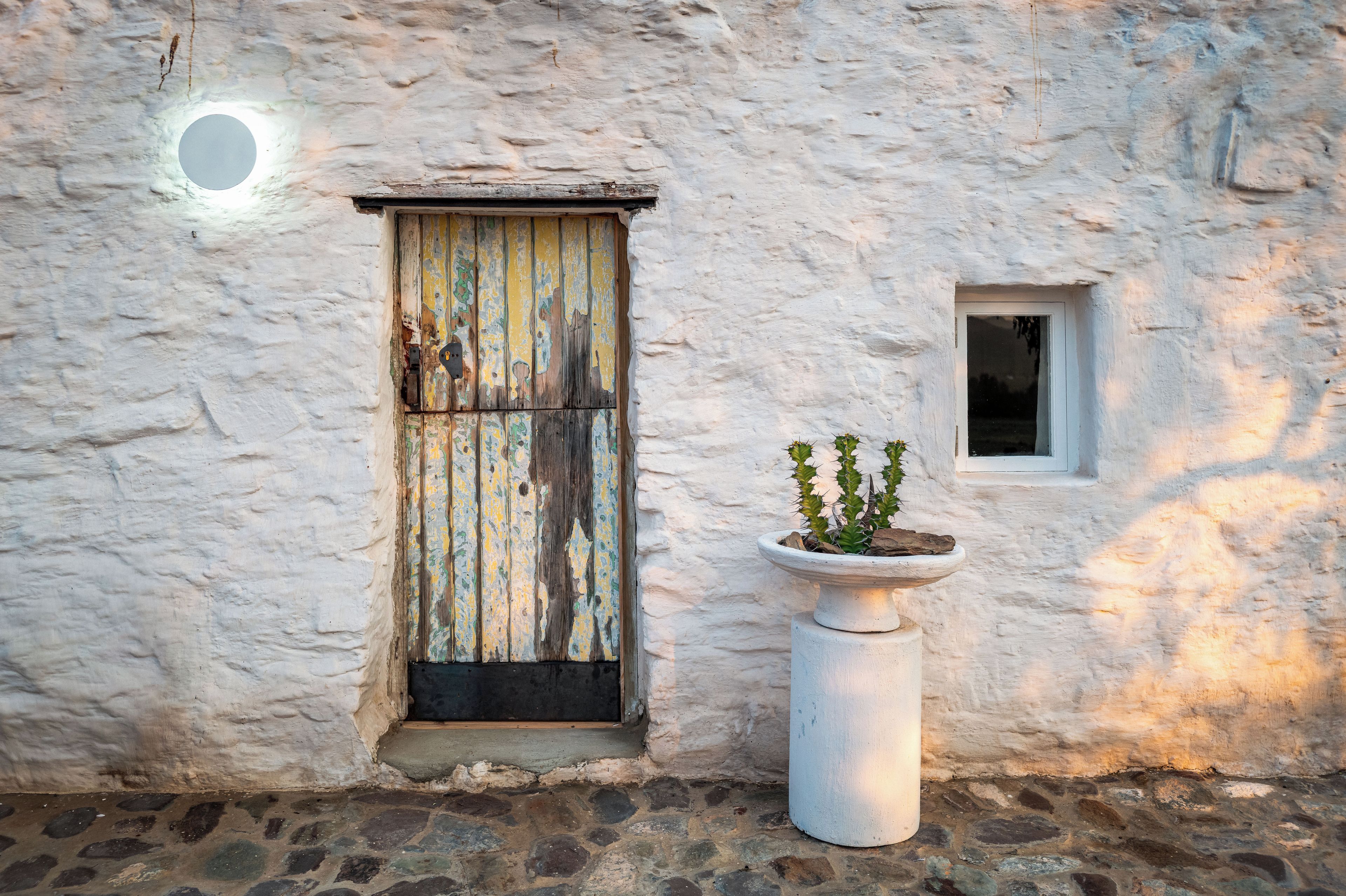 Un edificio rústico con una puerta de madera desgastada, un ventanal pequeño y una planta en maceta.