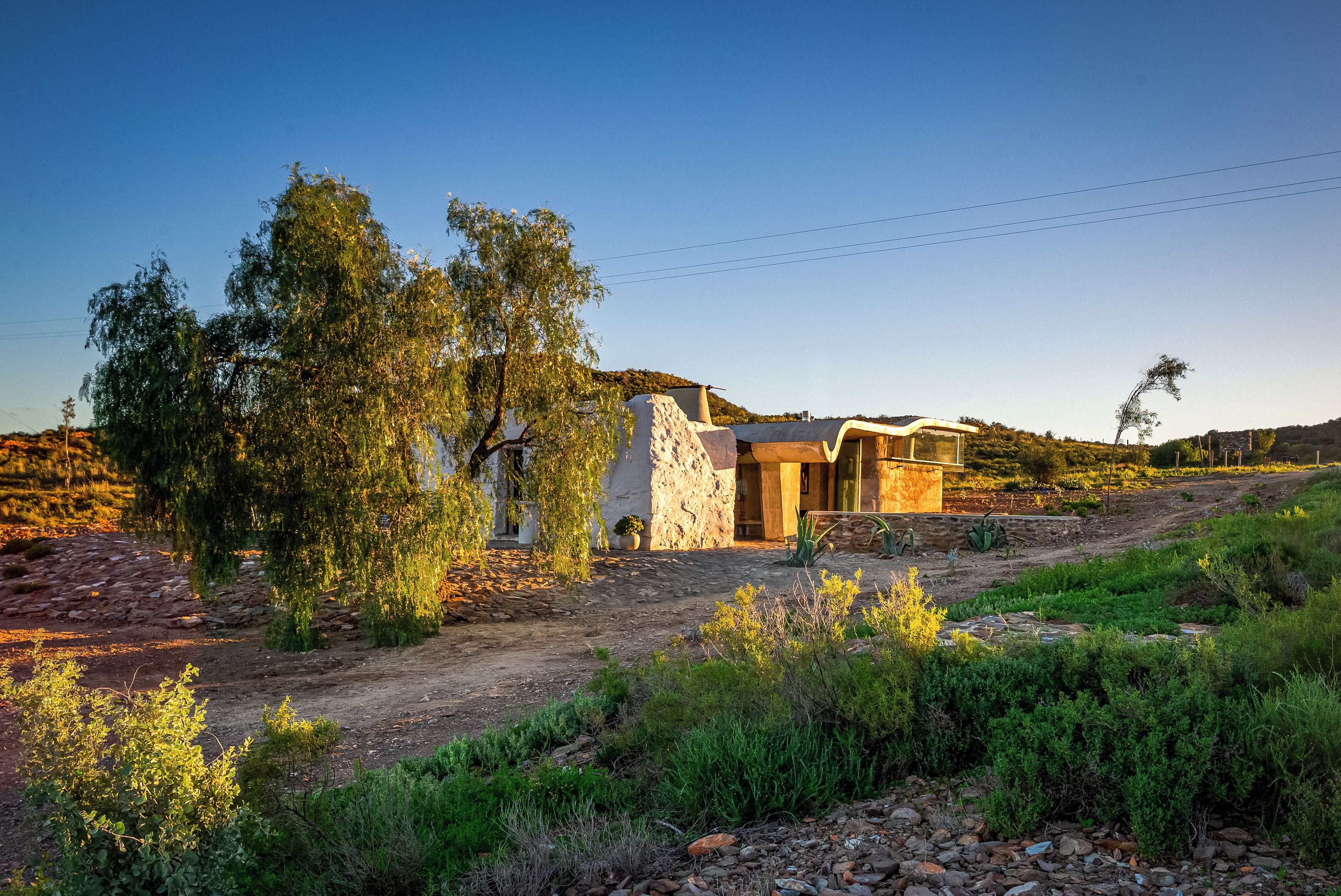 A rustic stone building with a modern extension, surrounded by natural vegetation and a clear sky.