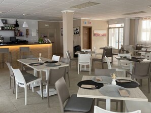 Ein modernes Restaurant mit weißen und grauen Tischen und Stühlen, einer Bar mit Flaschen und gerahmten Bildern an den Wänden.
